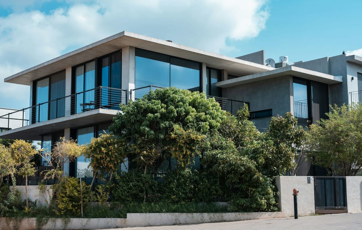 Casa moderna de concreto com janelas de vidro, varanda e jardim arborizado na frente