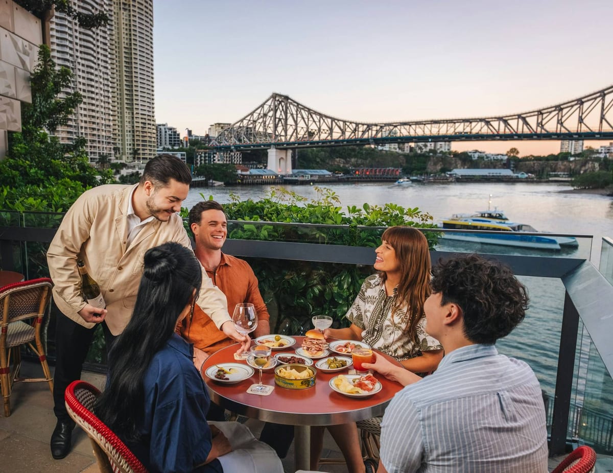 Amigos rindo à mesa de um terraço à beira do rio, com uma ponte ao fundo